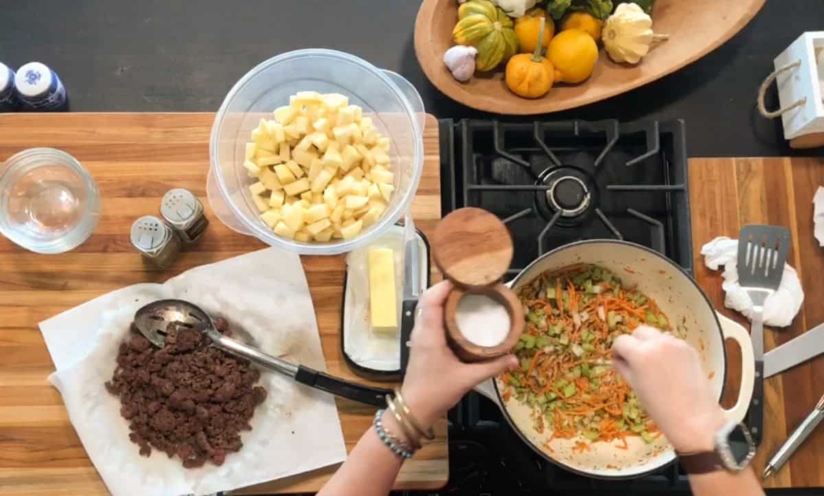 A person sprinkles salt into a pot of chopped vegetables cooking on a stovetop. Surrounding the pot are bowls of diced potatoes, cooked ground meat on paper towels, butter, and various squash on a wooden tray.