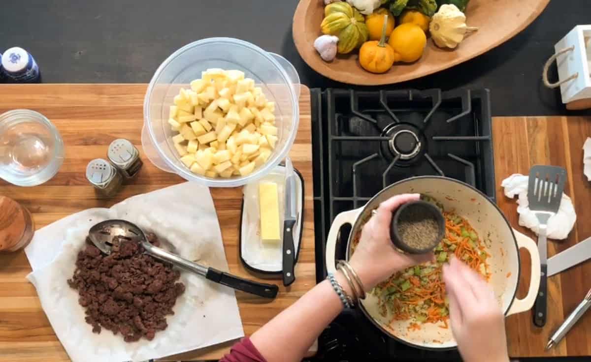 A person seasons shredded carrots and celery in a pot on a stove. Nearby, there are diced potatoes in a bowl, cooked ground beef on a plate, a stick of butter, and a tray of assorted squash.