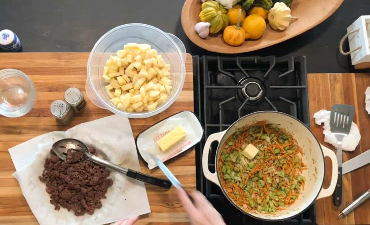 Overhead view of a kitchen counter with a pot of sautéed vegetables and butter on a stove, a pile of cooked ground meat, a bowl of diced potatoes, and various kitchen tools and ingredients nearby.