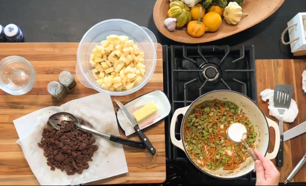 Overhead view of a kitchen counter with cooked ground beef, diced potatoes in a bowl, butter, seasonings, a glass of water, and a pot on the stove with sautéed vegetables being stirred with a spoonful of salt.