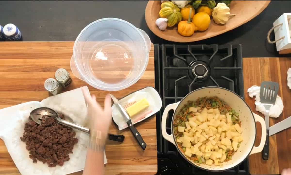 A person prepares food on a wooden countertop next to a stove. Cooked ground meat is on parchment paper, and chopped potatoes are in a pot with vegetables. Butter, knives, spices, and bowls are also visible. A gourd centerpiece is in the background.