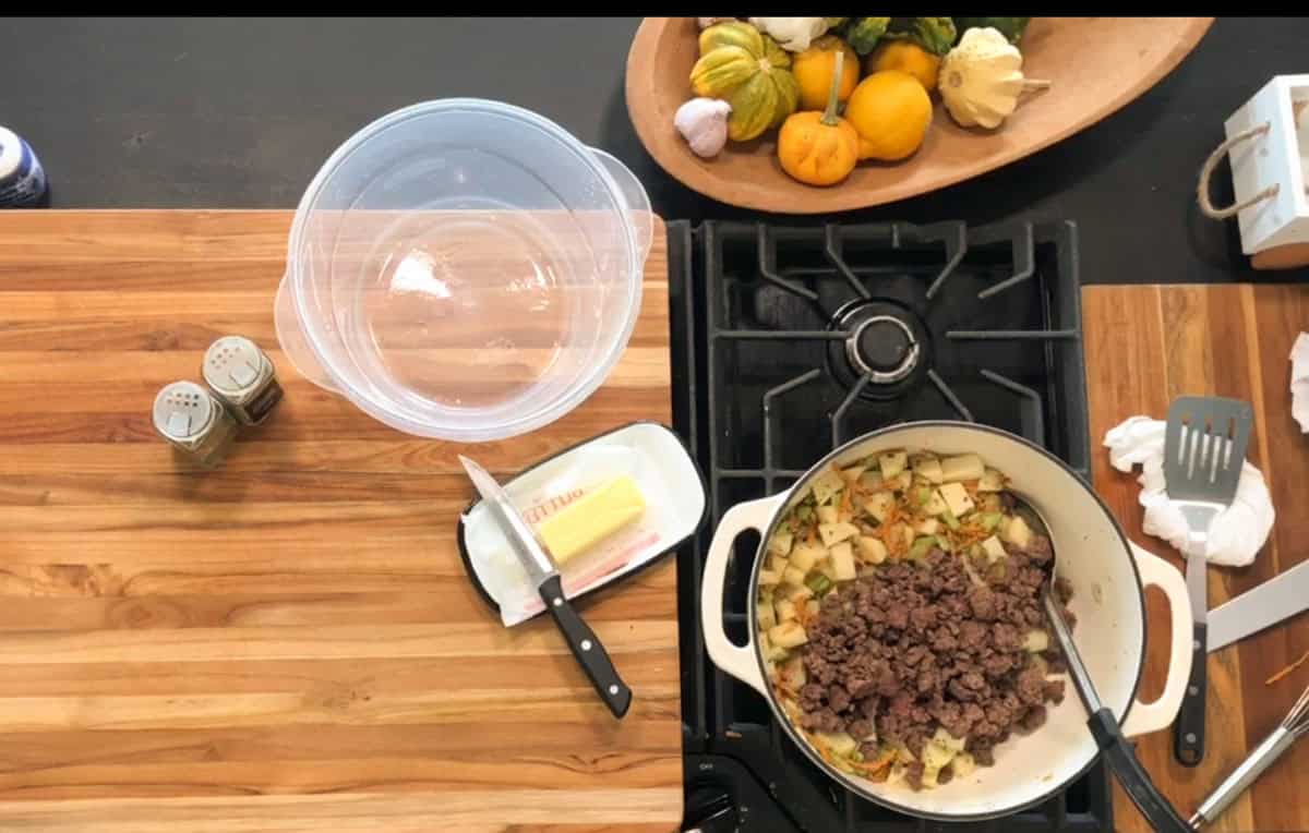 A stovetop scene shows a pot with cooked ground beef, diced potatoes, and vegetables. Nearby are butter, a knife, salt and pepper shakers, a plastic bowl, and a platter of assorted squash and garlic.