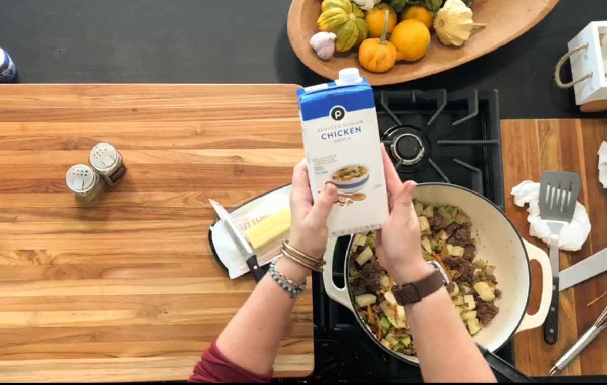 A person holds a carton of reduced sodium chicken broth over a pot on a stove, which contains chopped vegetables and meat. Nearby are seasonings, butter, a spatula, and a wooden bowl of assorted gourds.