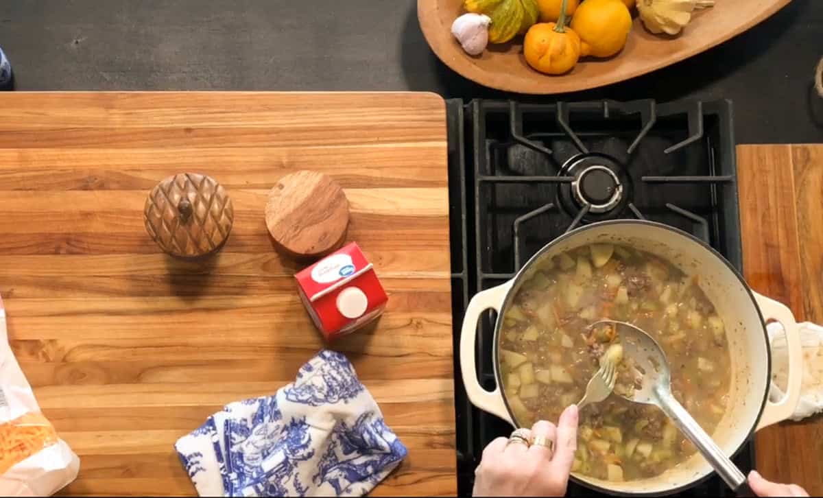 A person stirs a pot of soup with visible vegetables and meat on a stove. Next to the stove is a wooden cutting board holding a carton of cream, a wooden container, a bag, and a cloth. A bowl of gourds sits nearby.