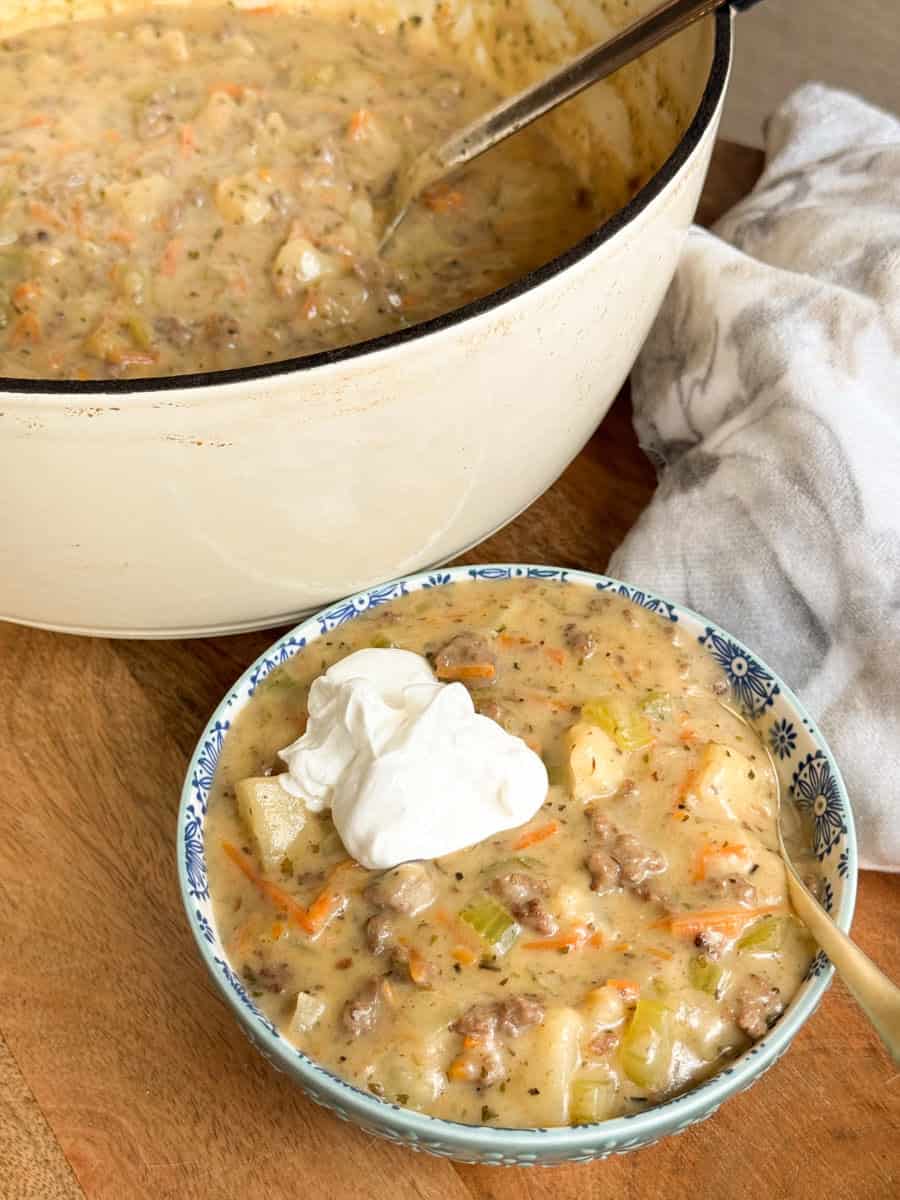 A bowl of creamy soup with ground meat, potatoes, carrots, and celery, topped with a dollop of sour cream, sits in front of a large pot filled with more of the same soup. A spoon and napkin are nearby.