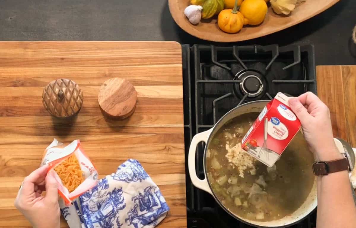 A person pours chicken broth from a carton into a pot of soup on a stove while holding a packet of dry pasta. Wooden salt and pepper containers and a decorative towel are on the counter next to the stove.