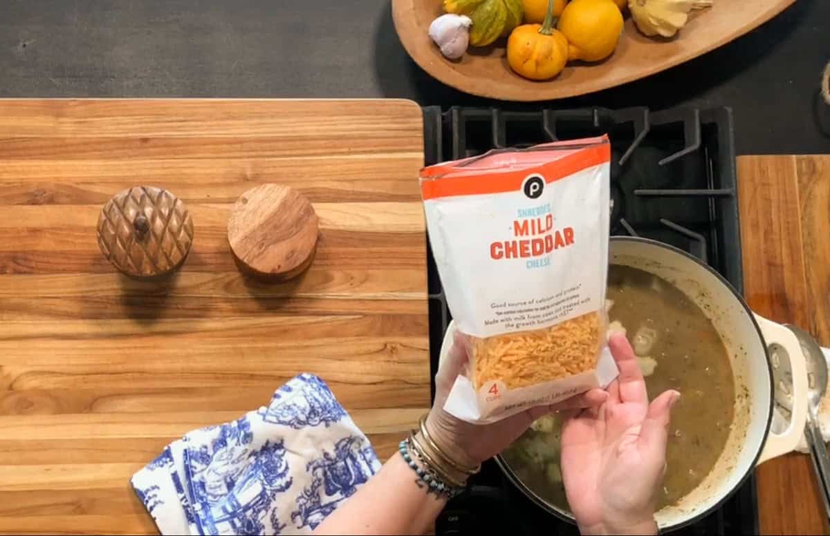 A person holds a bag of shredded mild cheddar cheese over a saucepan on a stovetop. A wooden cutting board, two wooden containers, and a decorative bowl of gourds and pumpkins are nearby.