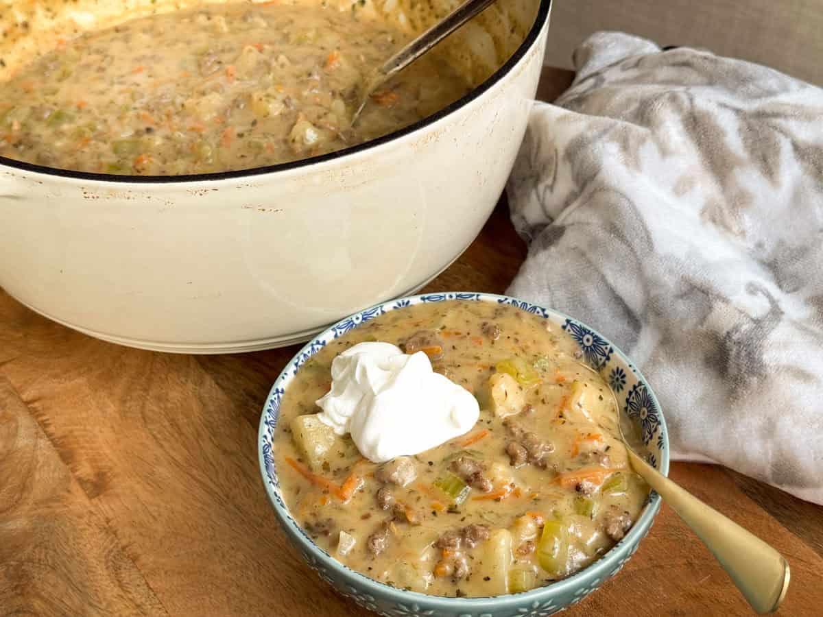 A bowl of creamy soup with vegetables, ground meat, and potatoes, topped with a dollop of sour cream, sits in front of a large white pot of the same soup on a wooden surface with a spoon and a cloth napkin nearby.