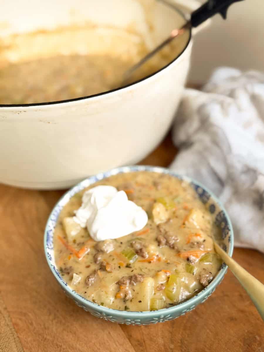 A bowl of creamy soup with ground meat, potatoes, carrots, and celery is topped with two dollops of sour cream. The bowl sits in front of a large pot of soup on a wooden surface with a spoon and a cloth nearby.