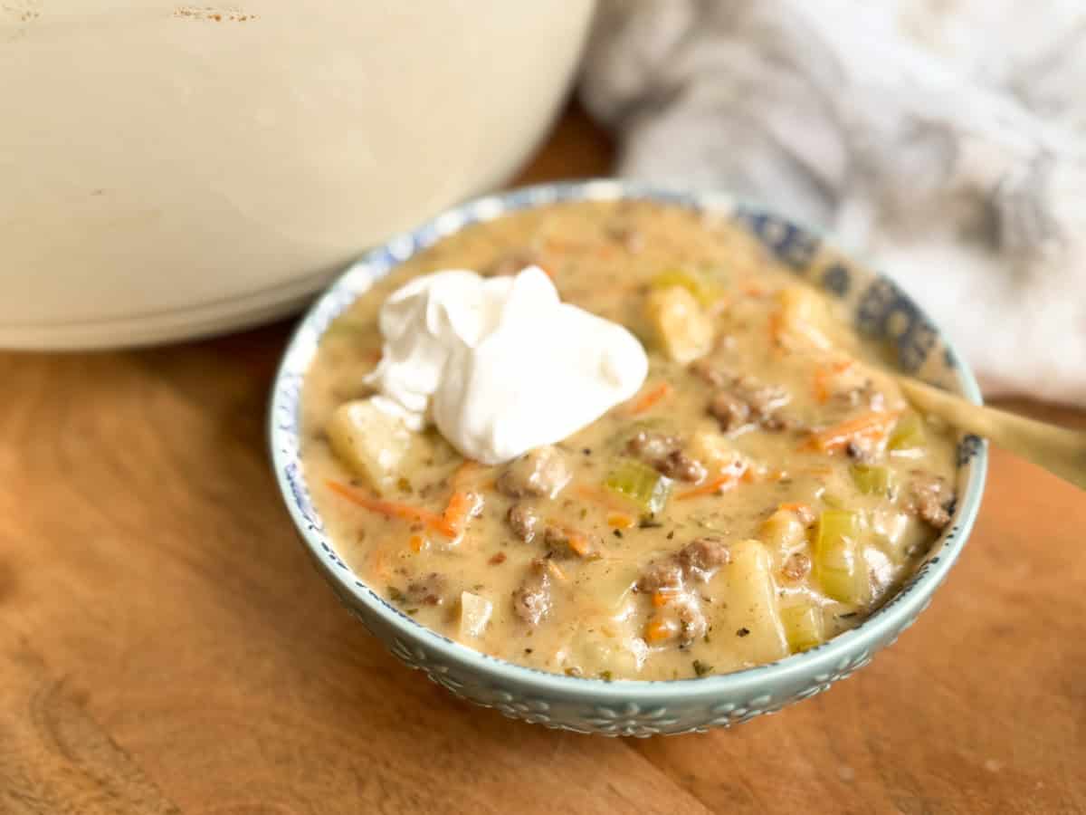 A bowl of creamy soup containing ground meat, potatoes, celery, and carrots, topped with a dollop of sour cream, sits on a wooden surface next to a white pot and a light-colored cloth.