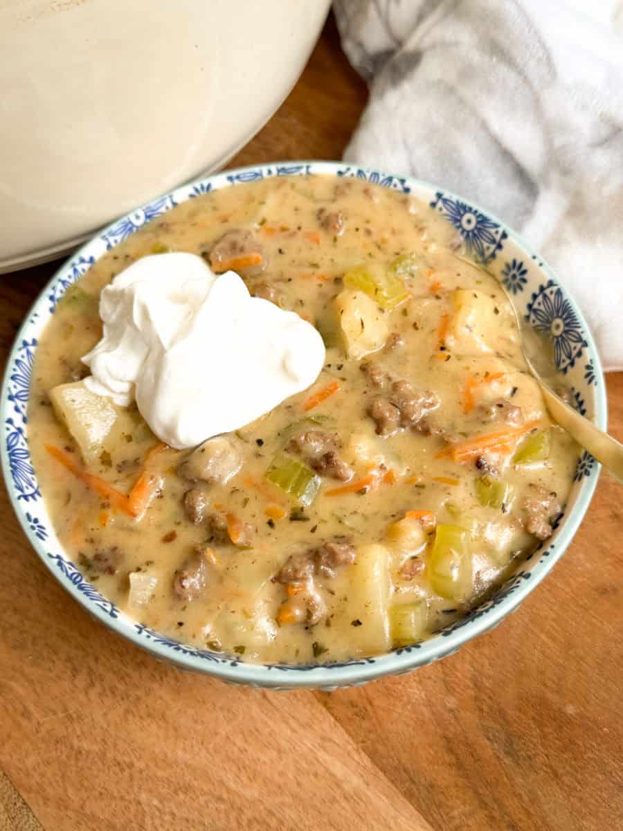 A bowl of creamy soup with ground meat, potatoes, carrots, and celery, garnished with a dollop of sour cream. A spoon rests in the bowl, which sits on a wooden surface next to a white cloth.