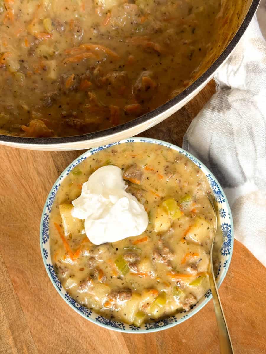 A bowl of creamy soup with ground meat, carrots, and celery, topped with a dollop of sour cream, sits on a wooden surface next to a pot of the same soup and a white kitchen towel.