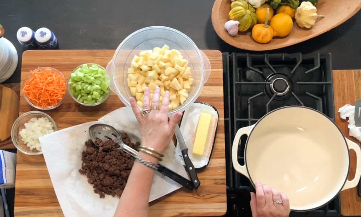 A person prepares to cook, with bowls of chopped potatoes, celery, onions, and carrots, cooked ground beef on a paper towel, a stick of butter, and a pot on the stove. Decorative squash sit in a bowl nearby.