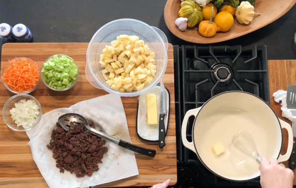 A person whisking butter in a white pot on a stove. On the counter are bowls of chopped potatoes, celery, carrots, onions, cooked ground beef, a knife, and a wooden tray with assorted squash.