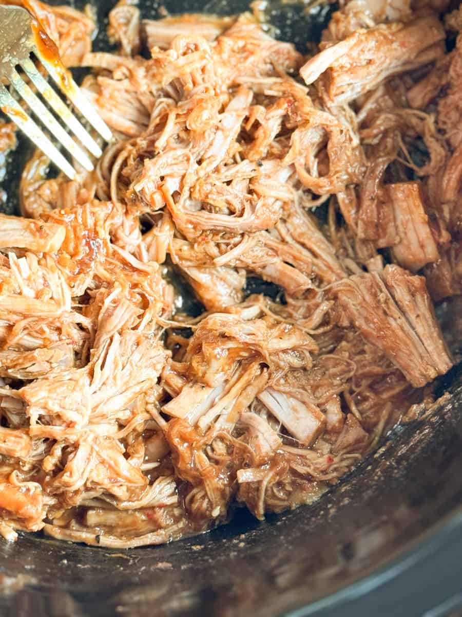 Shredded cooked meat mixed with sauce in a pan, with a metal fork visible on the side, pulling apart the meat. The meat appears tender and coated in a brown, glossy sauce.
