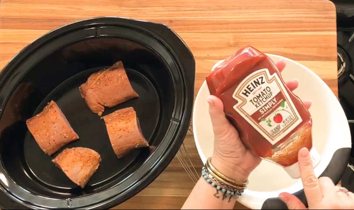 A hand holds a bottle of Heinz Tomato Ketchup above a white bowl. Nearby, a black slow cooker contains four pieces of seasoned pork on a wooden countertop.