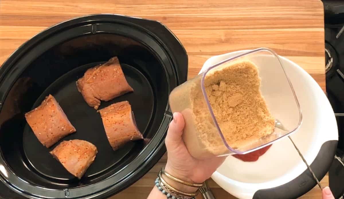 A person holds a container of brown sugar above a white bowl on a wooden countertop. Next to them is a slow cooker with four pieces of seasoned meat inside.