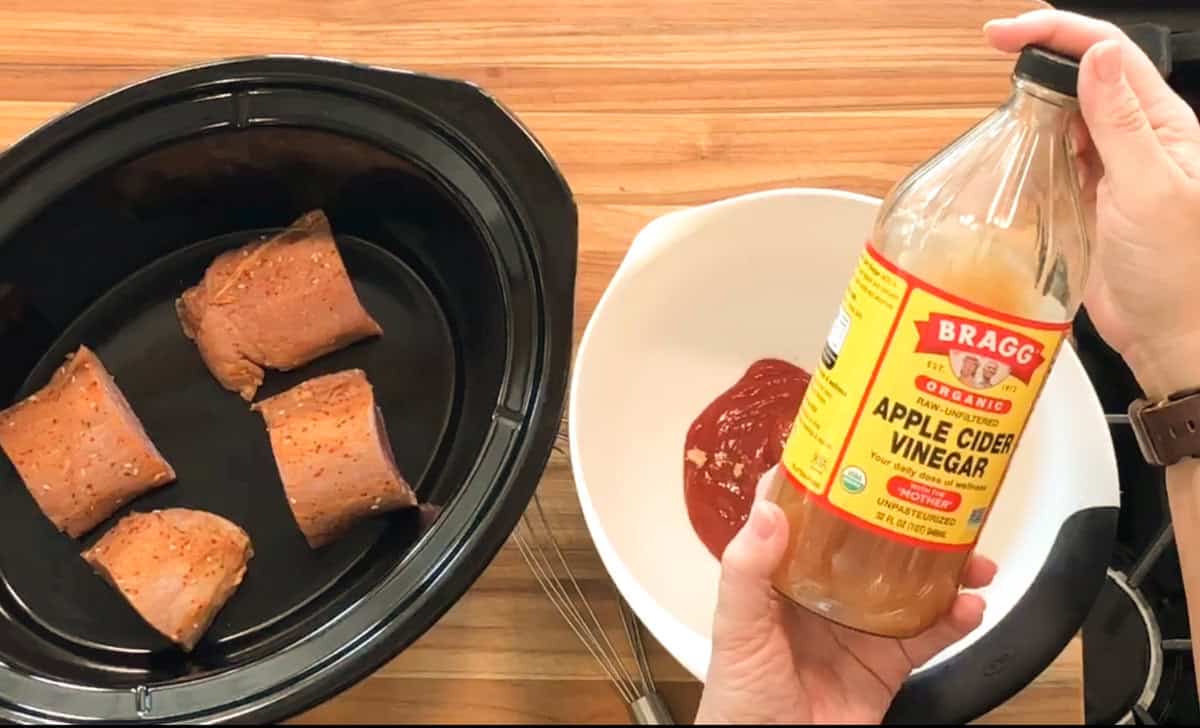 A person holds a bottle of Bragg Organic Apple Cider Vinegar above a bowl of ketchup next to a slow cooker with three pieces of seasoned meat. A whisk rests on the counter.