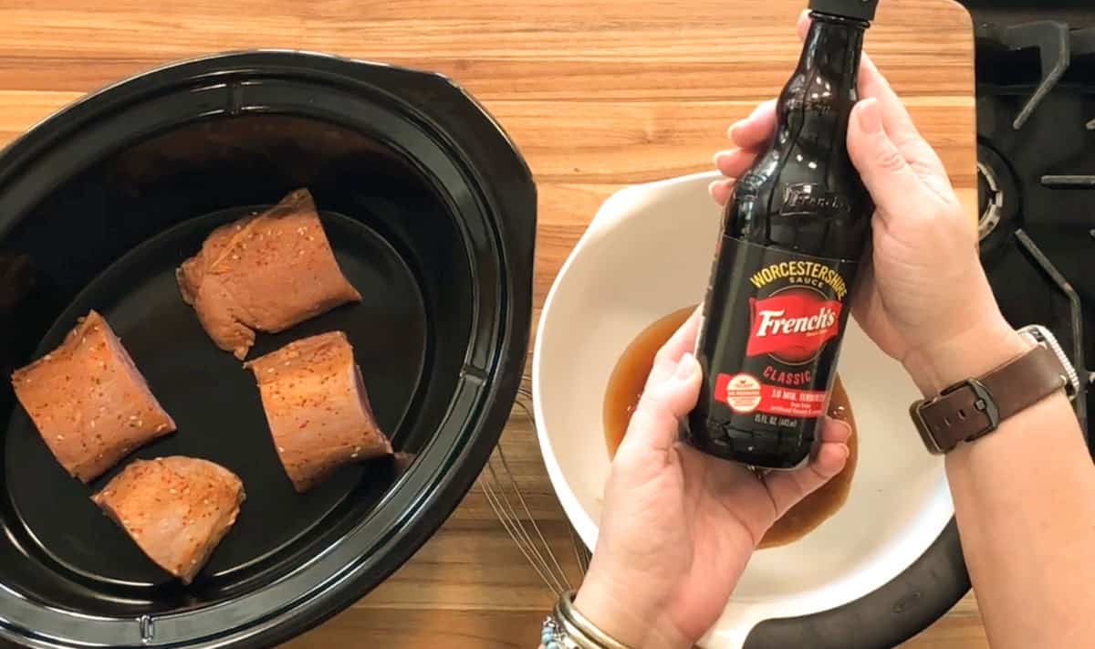 A person holds a bottle of French’s Worcestershire sauce above a white bowl with sauce. Next to it is a slow cooker containing three pieces of seasoned meat on a wooden surface.
