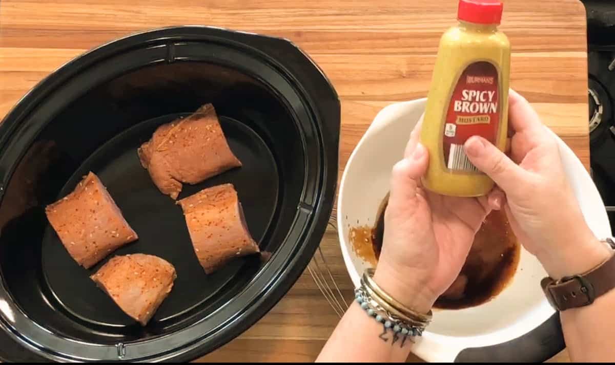 A person holds a bottle of spicy brown mustard above a bowl of sauce next to a slow cooker containing four pieces of seasoned meat, all on a wooden countertop.