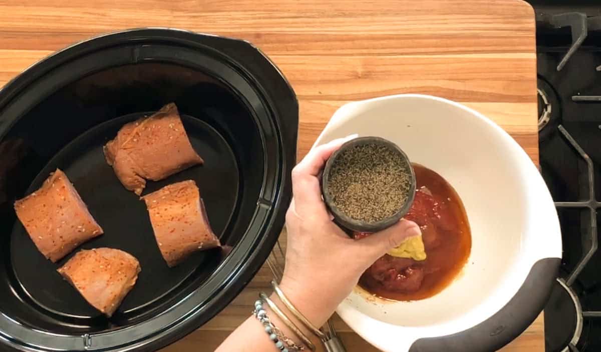A hand holds a container of dried herbs over a white bowl containing tomato sauce and mustard, next to a slow cooker with four pieces of raw seasoned meat on a wooden countertop.