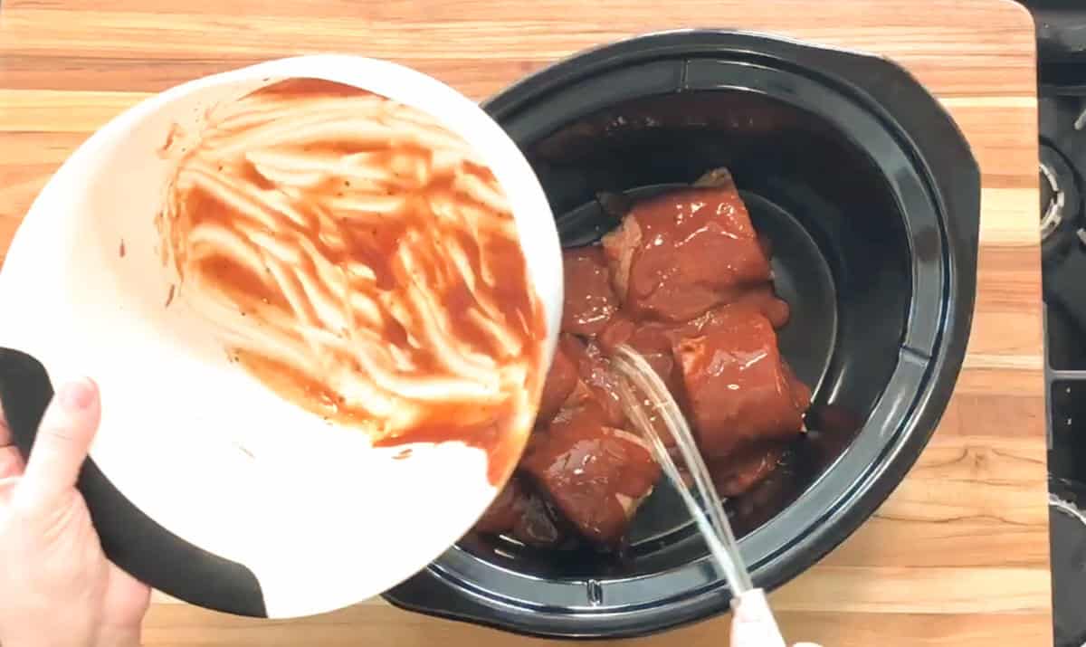 A hand pours sauce from a white bowl into a slow cooker containing pieces of raw, sauce-covered meat. The slow cooker sits on a wooden countertop.