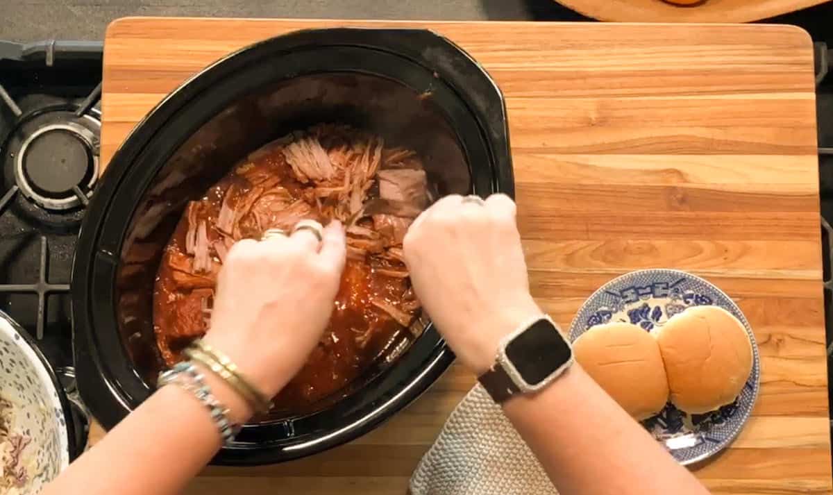 A person uses two forks to shred cooked meat in a slow cooker. Nearby, there is a plate with two sandwich buns and a folded cloth on a wooden countertop. The person is wearing bracelets and a smartwatch.