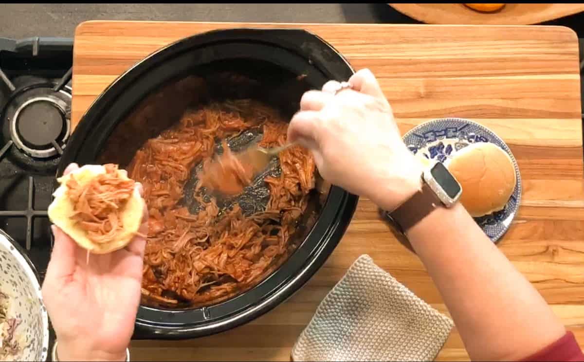 A person spoons shredded barbecue meat from a slow cooker onto a sandwich bun, with a plate holding another bun nearby on a wooden countertop.
