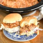 Two pulled pork sandwiches with coleslaw on round buns are on a blue and white plate. In the background, a slow cooker containing more pulled pork is visible on a wooden surface.