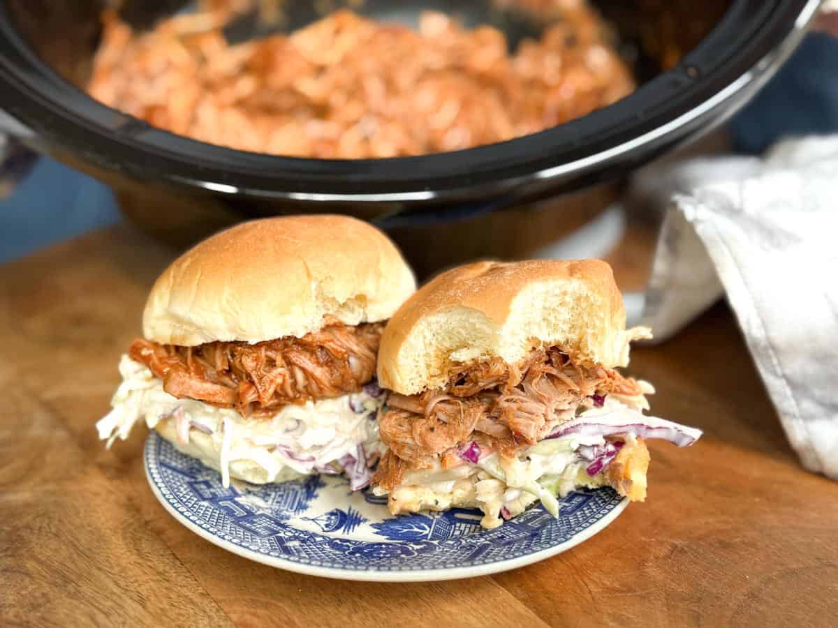 Two pulled pork sandwiches with coleslaw on a blue and white plate sit on a wooden surface. One sandwich has a bite taken out. In the background, a slow cooker filled with pulled pork is visible.