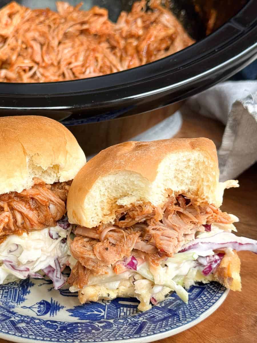Two pulled pork sandwiches with coleslaw on buns are served on a blue patterned plate. In the background, a slow cooker filled with more pulled pork is partially visible.