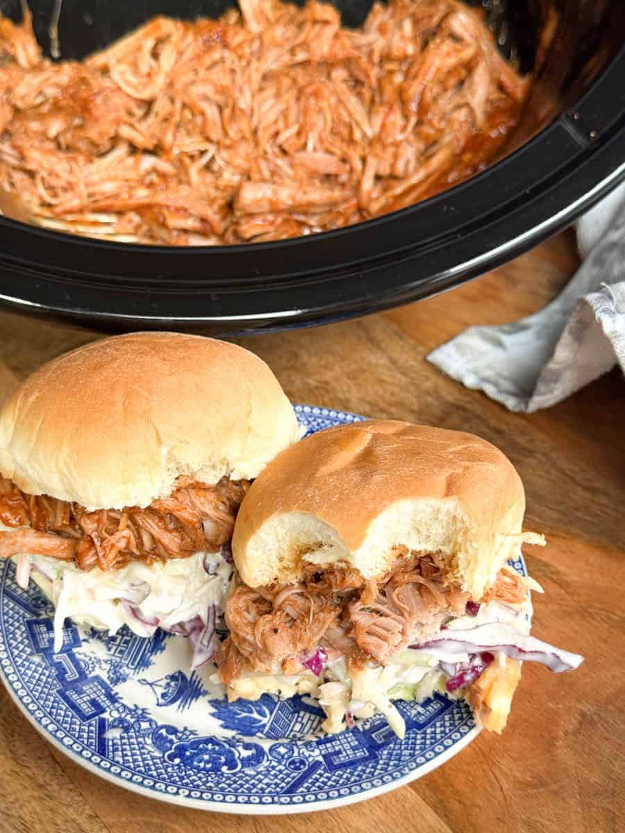 Two pulled pork sandwiches with coleslaw on a blue and white plate sit on a wooden surface. One sandwich has a bite taken out of it. In the background, there is a slow cooker filled with shredded pulled pork.
