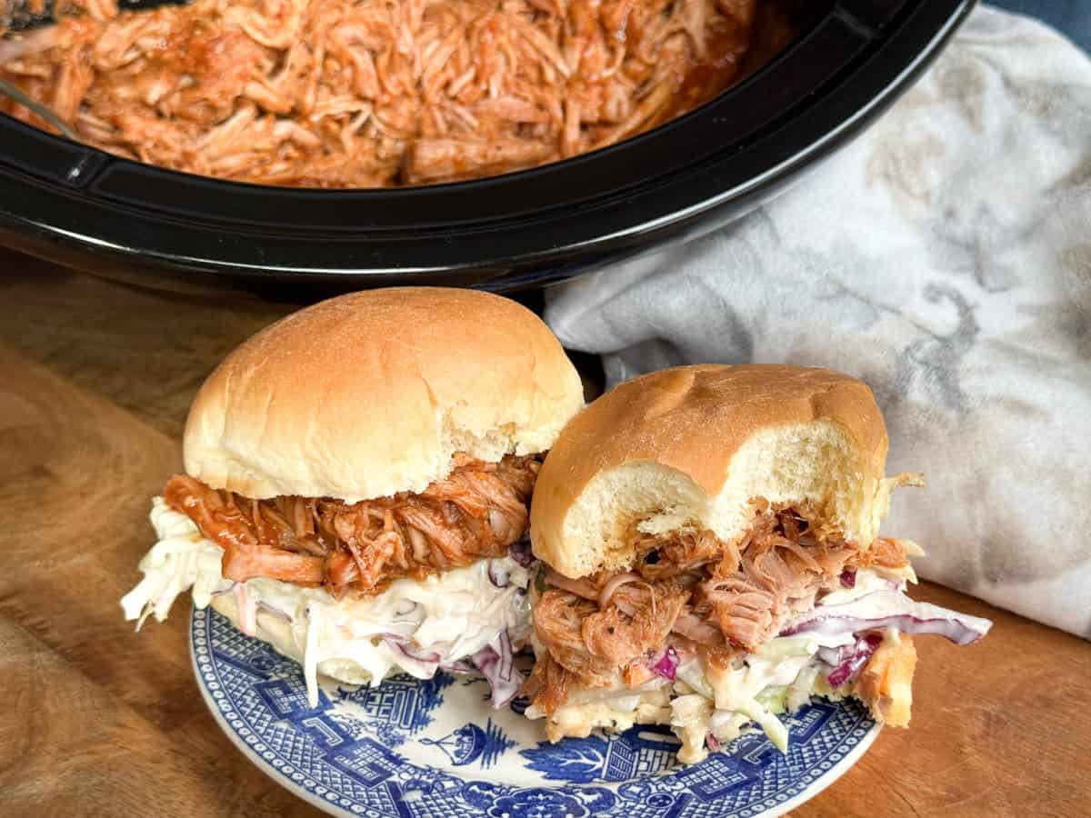Two pulled pork sandwiches with coleslaw on a blue and white plate. One sandwich has a bite taken out, and a slow cooker with more pulled pork is visible in the background.