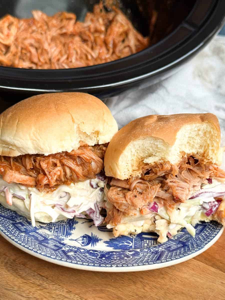 Two pulled pork sandwiches with coleslaw on a blue and white plate. One sandwich has a bite taken out. A slow cooker with shredded pulled pork is visible in the background.