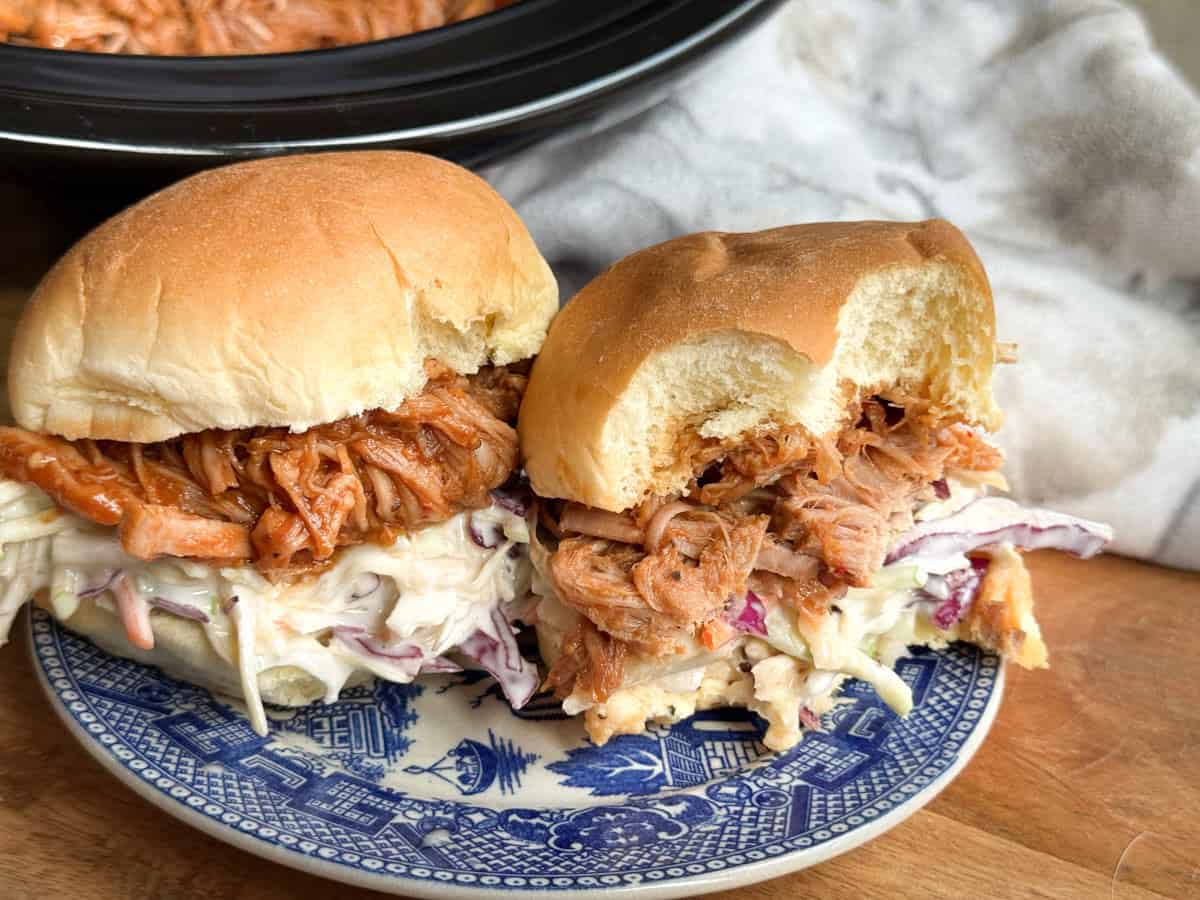 Two pulled pork sandwiches with coleslaw served on a blue and white patterned plate, placed on a wooden surface with a cloth and a slow cooker in the background.
