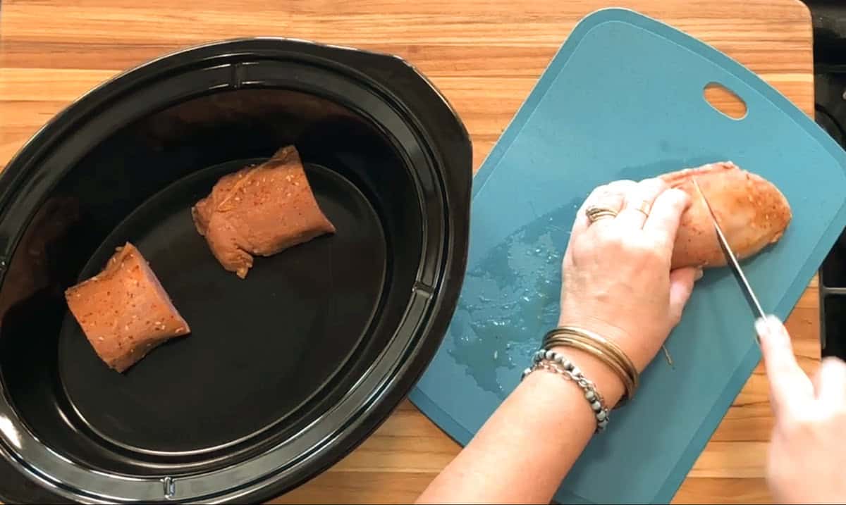A person slices seasoned pork tenderloin on a blue cutting board, placing pieces into a black slow cooker on a wooden countertop.