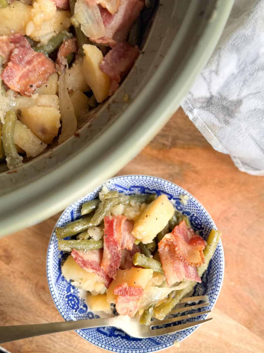 A bowl filled with green beans, potatoes, onions, and bacon slices sits on a wooden surface next to a slow cooker containing the same dish. A fork rests in the bowl.