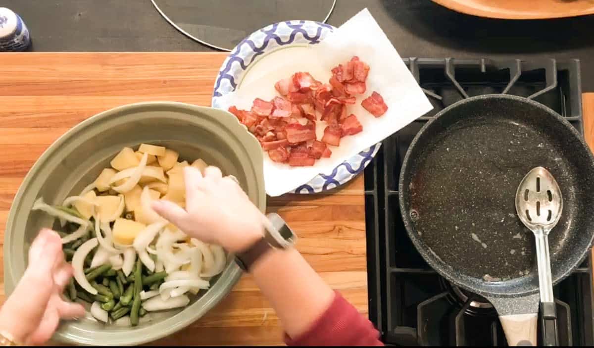 A person mixes sliced potatoes, onions, and green beans in a bowl on a wooden counter. Cooked bacon sits on a paper towel-lined plate nearby, and an empty pan with a slotted spoon rests on the stove.
