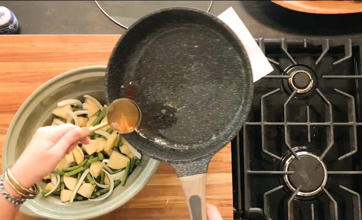 A person pours liquid from a frying pan with a ladle into a bowl containing sliced apples, green beans, and onions on a wooden countertop next to a gas stove.