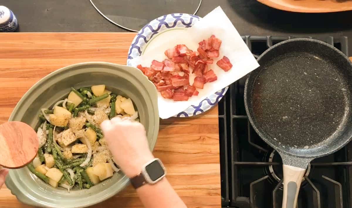 A person wearing a smartwatch adds seasoning to a slow cooker containing green beans and potatoes. Cooked bacon rests on a paper towel on a plate nearby; an empty frying pan is on the stovetop.