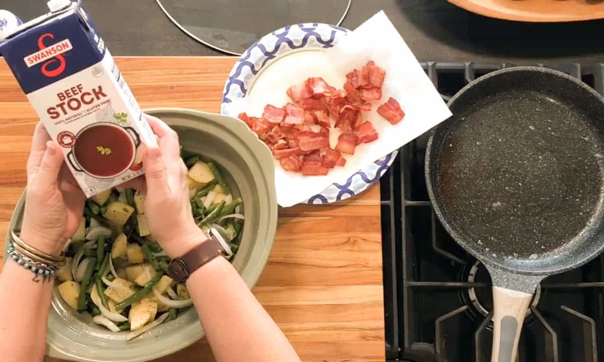 A person prepares to pour beef stock into a slow cooker containing potatoes, green beans, and onions. Cooked bacon pieces sit on a paper towel-lined plate next to a stove with a frying pan.
