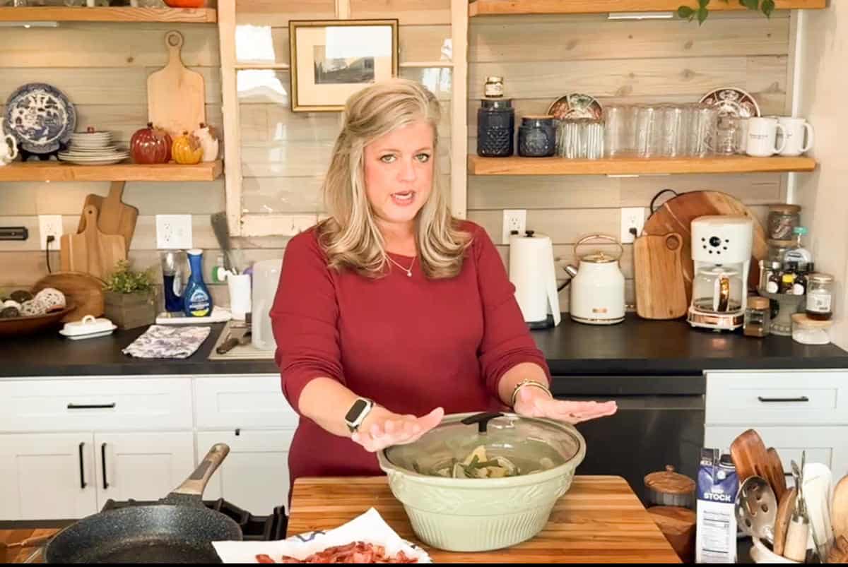 A woman with shoulder-length blonde hair stands in a kitchen behind a wooden counter, holding her hands over a large green mixing bowl. Various kitchenware and ingredients are visible on the shelves and counter around her.