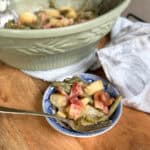 A small bowl filled with green beans, potatoes, onions, and bacon sits in front of a larger green serving dish containing more of the same food. A fork rests in the bowl, and a cloth napkin lies nearby on the wooden surface.