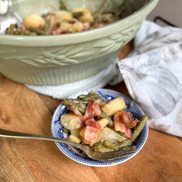 A small bowl filled with green beans, potatoes, onions, and bacon sits in front of a larger green serving dish containing more of the same food. A fork rests in the bowl, and a cloth napkin lies nearby on the wooden surface.