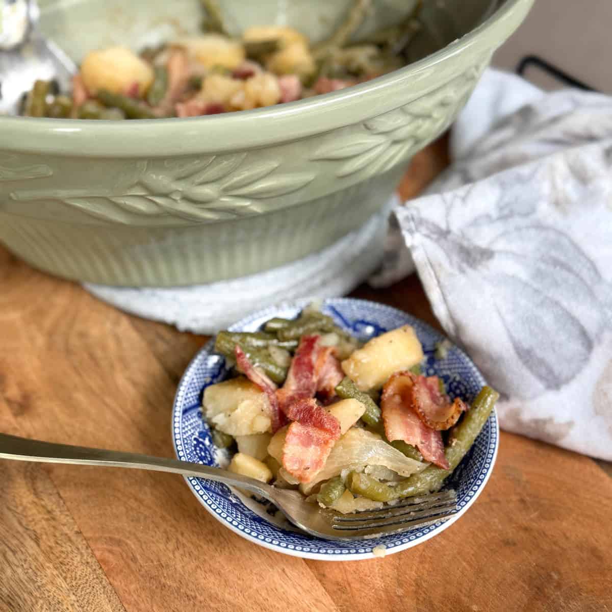 A small bowl filled with green beans, potatoes, onions, and bacon sits in front of a larger green serving dish containing more of the same food. A fork rests in the bowl, and a cloth napkin lies nearby on the wooden surface.