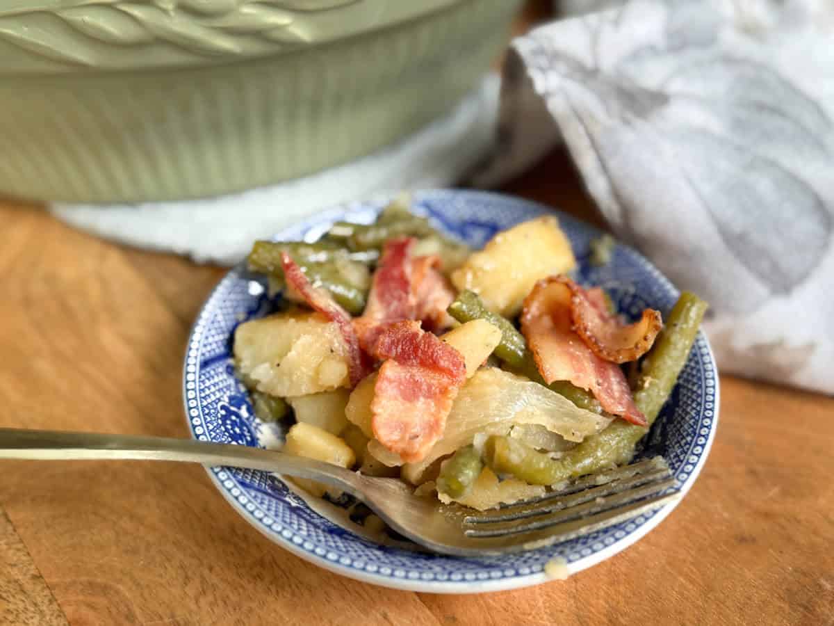 A small blue and white dish containing cooked green beans, potatoes, onions, and pieces of bacon sits on a wooden surface next to a fork, with a green bowl and white cloth in the background.