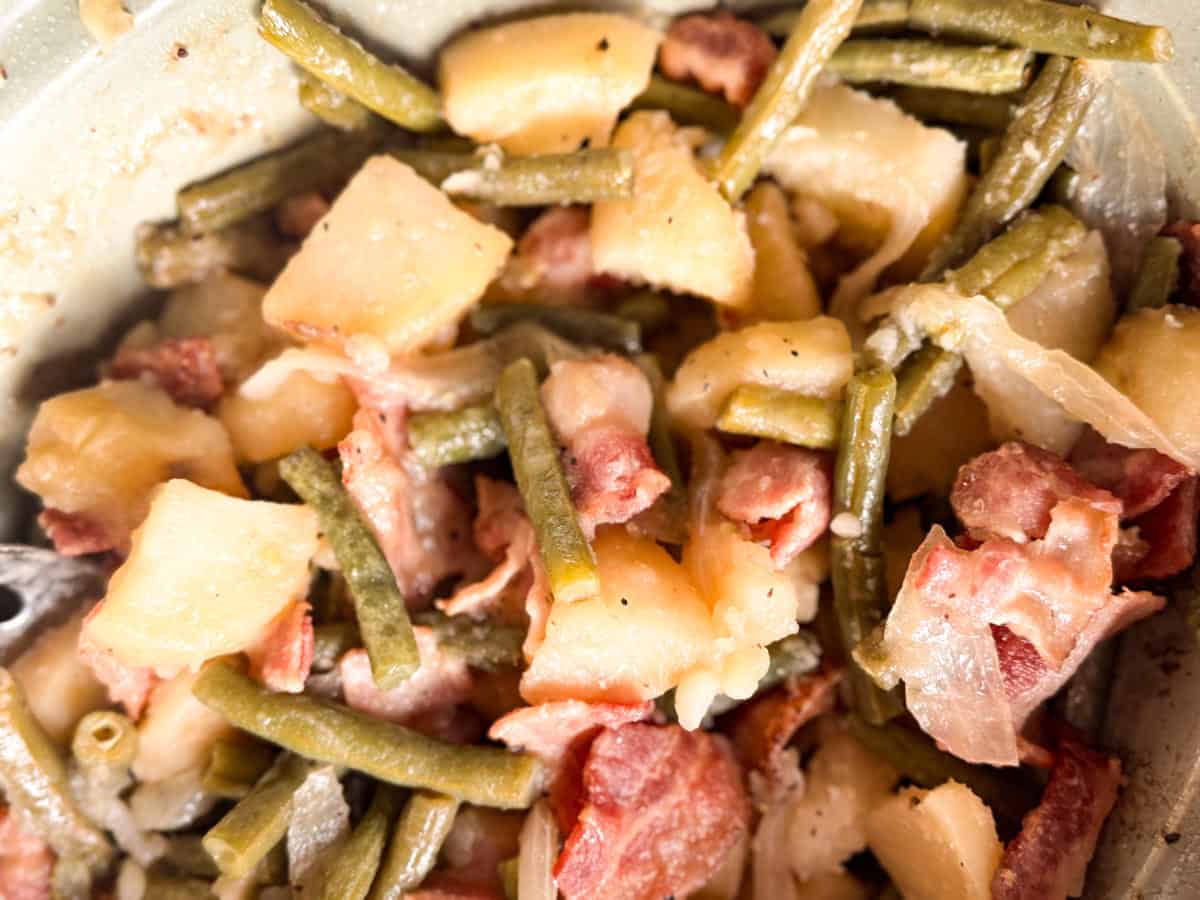 A close-up of a dish containing green beans, diced potatoes, bacon pieces, and onions, all mixed together and cooked. The ingredients appear soft and seasoned.