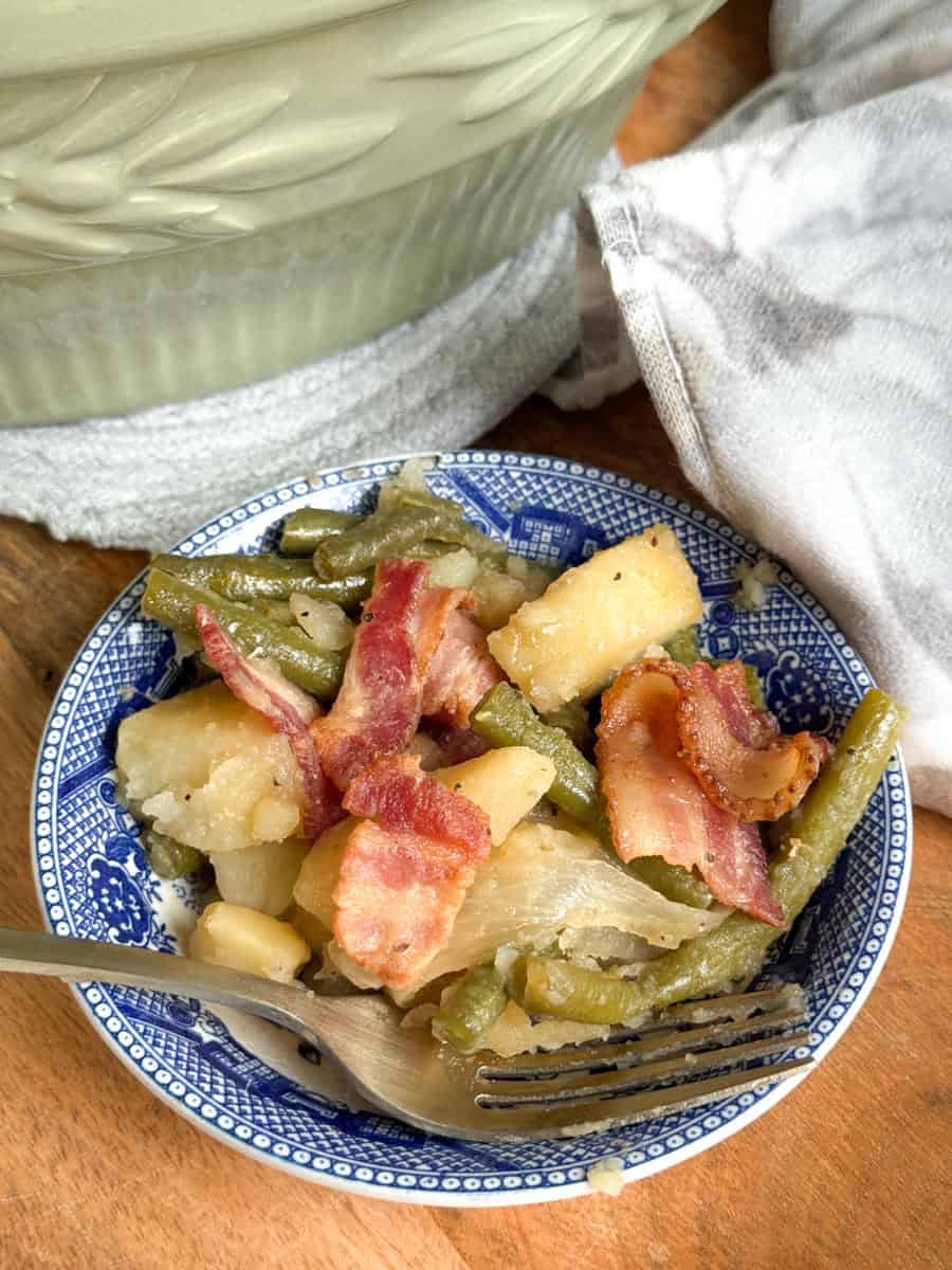 A blue and white plate holds a serving of green beans, potatoes, onions, and bacon slices. A fork rests on the plate, and part of a green dish and white towel are visible in the background.