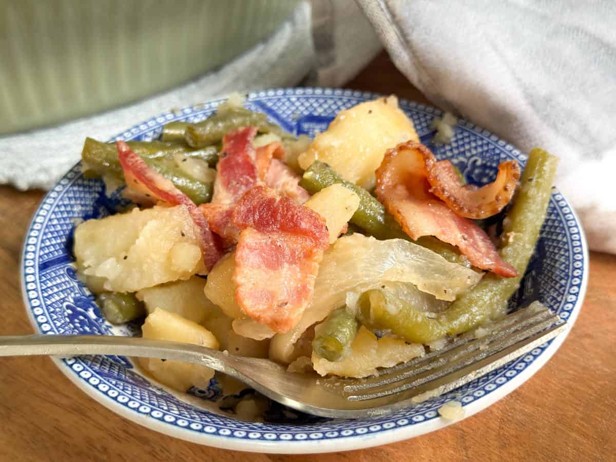 A blue and white patterned plate holds a serving of sliced potatoes, green beans, onions, and crispy bacon, with a fork resting on the side. A white cloth napkin and a green dish are in the background.