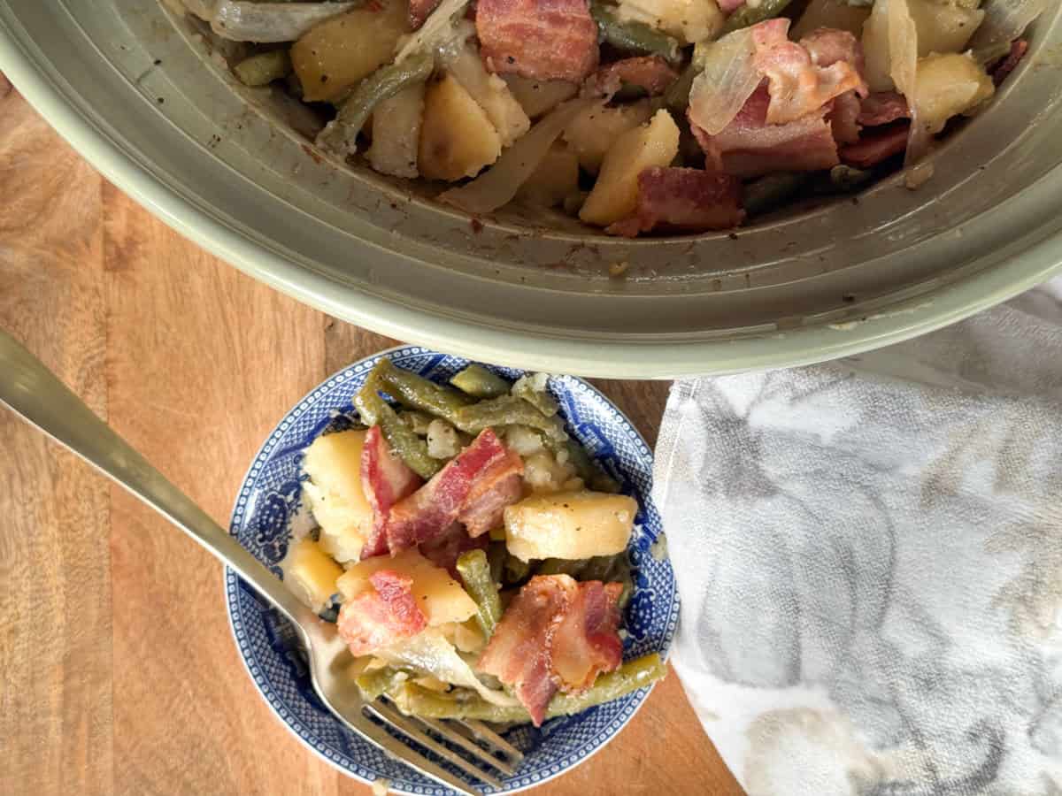 A bowl filled with a mixture of green beans, potatoes, onions, and bacon sits on a wooden surface next to a slow cooker containing more of the same dish. A fork rests in the bowl and a cloth napkin is nearby.